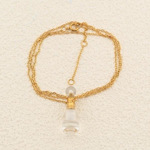 May include: A gold-colored necklace with a small, clear glass perfume bottle pendant. The bottle has a gold-colored cap and a clear stopper. The necklace chain is delicate and has a simple clasp.