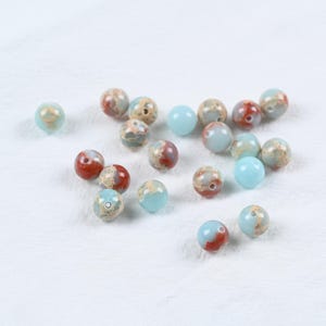 May include: A selection of round beads in shades of light blue, red, and tan. Some beads feature a speckled design, while others are plain. The beads are arranged on a white background.