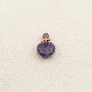 May include: A heart-shaped purple amethyst perfume bottle pendant featuring a polished stone body, a gold-toned metal collar, and a small round purple amethyst stopper. The piece is designed with a smooth, glossy finish and a deep violet hue.