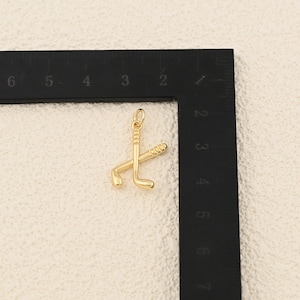May include: A gold-colored pendant featuring two crossed golf clubs. The pendant has a small loop at the top for attaching to a chain. The golf clubs are detailed with grooves and curves, suggesting a miniature sports-themed accessory.