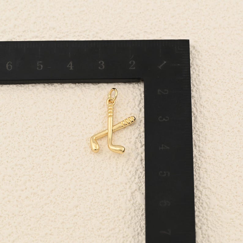 May include: A gold-colored pendant featuring two crossed golf clubs. The pendant has a small loop at the top for attaching to a chain. The golf clubs are detailed with grooves and curves, suggesting a miniature sports-themed accessory.