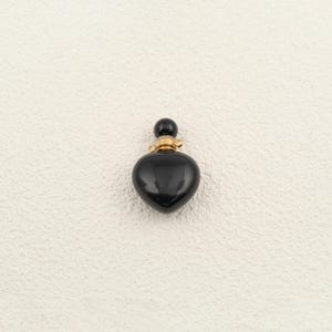 Puede incluir: Un pequeño colgante negro en forma de corazón con un acabado liso y brillante. La parte superior del corazón presenta una tapa de metal dorado con una pequeña cuenta negra redonda y una diminuta anilla incorporada para colgarlo.