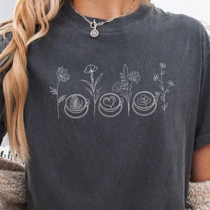 Close up of a model wearing a Pepper dark grey Comfort Colors 1717 t-shirt with a retro Coffee and Flowers graphic. Showcasing a trendy cozy barista aesthetic and soft cotton texture.