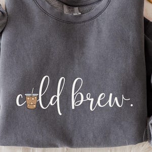Könnte beinhalten: Ein anthrazitfarbenes Sweatshirt mit Rundhalsausschnitt und dem weißen Schreibschrift-Text "cold brew.", wobei ein Becher mit Eiskaffee den Buchstaben "o" ersetzt. Das Kleidungsstück hat lange Ärmel sowie gerippte Bündchen am Kragen und Saum.