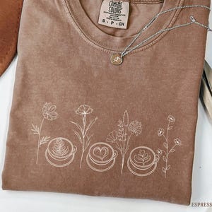 May include: Espresso-colored t-shirt with a crew neck and short sleeves. The shirt features a white floral design with coffee cups. A silver necklace with a small pendant is draped across the shirt. The tag reads "Comfort Colors".