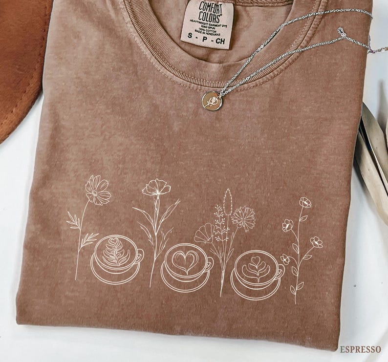 May include: Espresso-colored t-shirt with a crew neck and short sleeves. The shirt features a white floral design with coffee cups. A silver necklace with a small pendant is draped across the shirt. The tag reads "Comfort Colors".