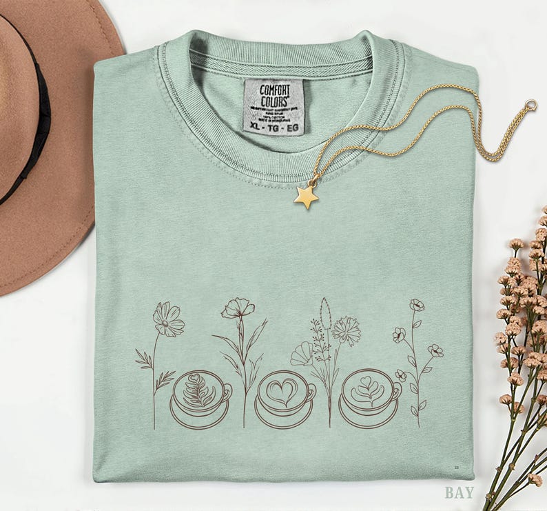 May include: A light green t-shirt with a floral and coffee cup design. The shirt features a gold star necklace and a tag that reads "COMFORT COLORS" and "XL - TG - EG". The design includes three coffee cups with latte art and various flowers.