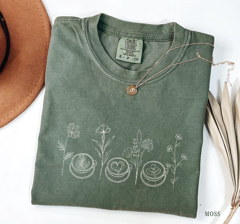 May include: A moss green t-shirt with a white floral and coffee cup design. The shirt has a crew neck and short sleeves. A gold necklace with a small pendant rests on the shirt. The shirt is labeled "Comfort Colors" and is size small.
