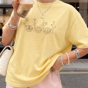 May include: A light yellow t-shirt featuring a black line drawing of coffee art and flowers. The shirt is a soft, casual style. The person is wearing bracelets and a necklace with a moon pendant.