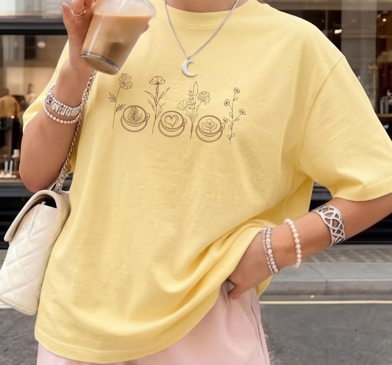 May include: A light yellow t-shirt featuring a black line drawing of coffee art and flowers. The shirt is a soft, casual style. The person is wearing bracelets and a necklace with a moon pendant.