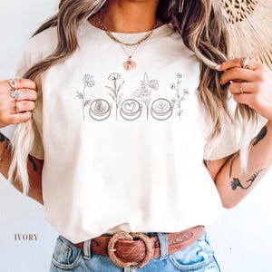 May include: Ivory-colored t-shirt featuring a line drawing of coffee cups with latte art and floral elements. The text "IVORY" is printed at the bottom of the shirt. The shirt is paired with blue jeans and a brown belt.