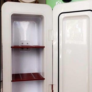 May include: A white mini-fridge with an open door, featuring a red and white emblem on top. The interior has two red shelves. The fridge is designed with a retro aesthetic.