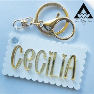 May include: A personalized keychain with the name "Cecilia" in gold lettering on a white, scalloped-edge tag. The keychain has a gold clasp and ring, with a small clear charm attached. The logo "The Artsy Trio" is in the upper right corner.