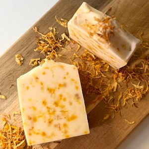 Può includere: Due saponette artigianali beige chiaro con macchie arancioni, su un tagliere di legno. Il sapone è decorato con petali d'arancia essiccati. Lo sfondo è bianco. Le saponette misurano circa 8 cm di lunghezza.