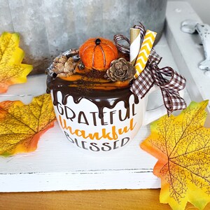 Puede incluir: Una taza de cer&aacute;mica blanca con el texto GRATEFUL thankful BLESSED en negro y naranja. La taza est&aacute; decorada con goteo de chocolate falso, una calabaza de fieltro, dos pi&ntilde;as, dos pajitas de papel y un lazo de cuadros marr&oacute;n y blanco.