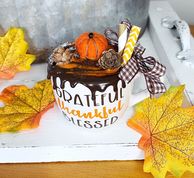 Puede incluir: Una taza de cer&aacute;mica blanca con el texto GRATEFUL thankful BLESSED en negro y naranja. La taza est&aacute; decorada con goteo de chocolate falso, una calabaza de fieltro, dos pi&ntilde;as, dos pajitas de papel y un lazo de cuadros marr&oacute;n y blanco.