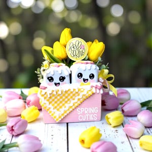 May include: A pink wooden box with the words "Spring Cuties" holds two marshmallow characters with smiling faces, surrounded by yellow tulips and pink and yellow tulips. A yellow and white gingham cloth is visible. A circular sign says "Hello Spring".