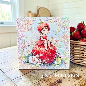 May include: A square 3 x 3 inch mini sign featuring a whimsical illustration of a small fairy with wings wearing a strawberry hat, sitting atop a large red strawberry surrounded by white flowers, all accented with a sparkling glitter finish.