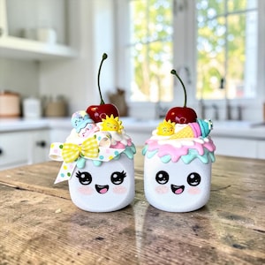 May include: Two white, smiling, decorative jars with ice cream-themed toppers. Each jar has a cherry on top, with one featuring a yellow bow and the other a small ice cream cone. The jars are on a wooden table.