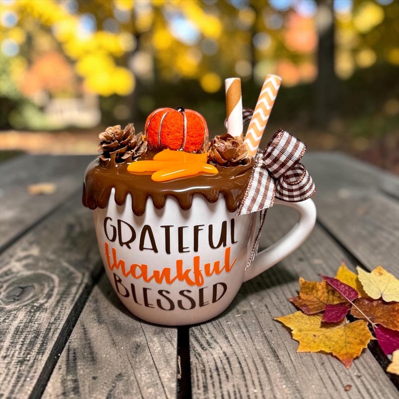 May include: A white ceramic mug featuring the text "GRATEFUL thankful BLESSED" in brown and orange. The top is decorated with brown faux-drip icing, a small orange pumpkin, pinecones, two striped paper straws, and a brown and white gingham bow.