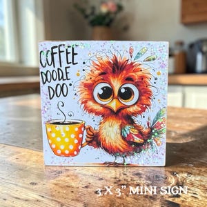 May include: A square 3 x 3 inch wooden sign featuring a colorful illustration of a fluffy orange bird holding a yellow polka-dot coffee mug. The text COFFEE DOODLE DOO is printed in black on the left side, with iridescent glitter accents throughout.