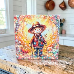 May include: A square mini sign measuring 3 x 3 inches featuring a colorful illustration of a smiling scarecrow wearing a hat and scarf, standing in an autumn landscape with pumpkins and flowers, accented with iridescent glitter throughout the design.