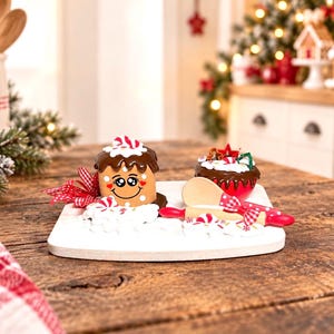 May include: A decorative gingerbread-style figure with a smiling face, brown icing, and a red-and-white bow, displayed on a white board alongside a miniature chocolate-topped cupcake, a small wooden spoon, and a red-handled rolling pin.
