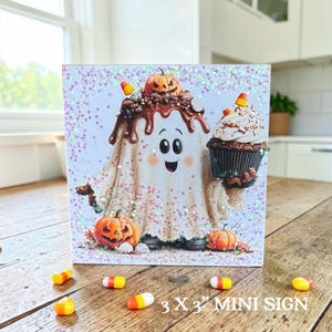 May include: A square sign featuring a cartoon ghost covered in chocolate frosting and sprinkles, holding a cupcake. The ghost is flanked by two orange pumpkins, with candy corn scattered around the base. The text reads 3 X 3 MINI SIGN.