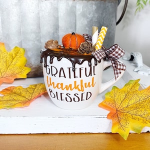 Puede incluir: Una taza de cer&aacute;mica blanca con el texto GRATEFUL thankful BLESSED en marr&oacute;n y naranja. La taza est&aacute; decorada con falso sirope de chocolate, una calabaza de fieltro, pi&ntilde;as, pajitas de papel y un lazo de cinta a cuadros marr&oacute;n y blanco.