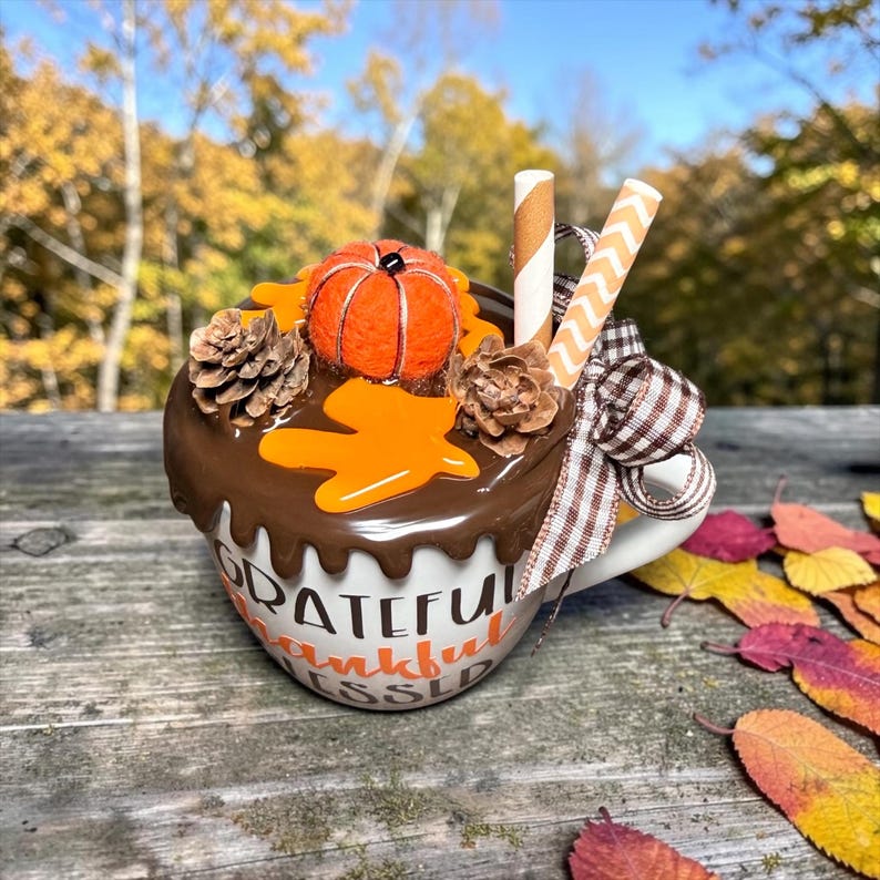 May include: A white ceramic mug featuring the text Grateful Thankful Blessed in black and orange lettering. The top is decorated with brown faux icing, a small orange fabric pumpkin, two pinecones, two paper straws, and a brown plaid ribbon.