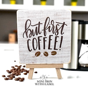 May include: A small, square sign on a wooden easel with the phrase "but first COFFEE!" in brown script. The sign has a textured, off-white background and three coffee bean illustrations. The sign is 4" x 4".