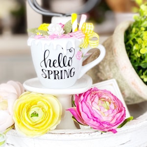 May include: A white ceramic mug with the words "hello SPRING" in black script, decorated with pink and yellow flowers, and a yellow gingham bow. The mug sits on a small white pedestal, surrounded by colorful flowers.