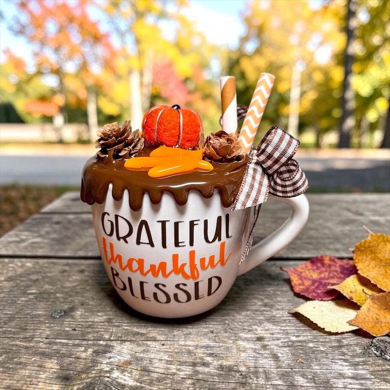 May include: A white ceramic mug featuring the text "GRATEFUL thankful BLESSED" in brown and orange. The top is decorated with brown faux-drip icing, a small orange pumpkin, pinecones, two paper straws, and a brown and white checkered ribbon bow.