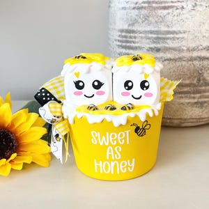 May include: A yellow ceramic mug with the text "Sweet as Honey" and two white bee-shaped marshmallows with black eyes and pink cheeks. The mug is decorated with yellow and black bees.