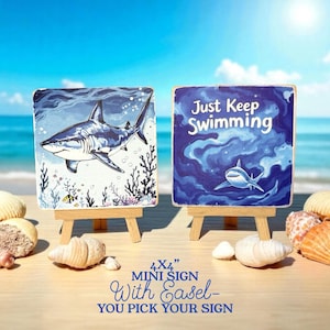 May include: Two 4x4 inch mini signs with easels. One sign features a shark illustration, the other says "Just Keep Swimming". The signs are on a wooden surface with seashells and the ocean in the background.