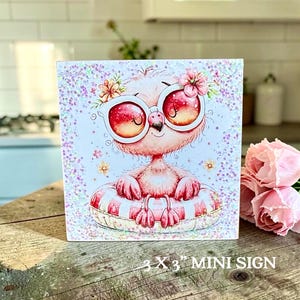 May include: A square 3 x 3 inch mini sign featuring a cartoon flamingo wearing large pink sunglasses and sitting in a striped pool float. The illustration is decorated with small flowers and iridescent glitter accents on a white background.