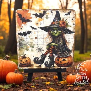 May include: A square 4x4 inch mini sign on a black easel featuring a green-skinned witch in a pointed hat, orange and black striped stockings, and black boots. The illustration includes black bats, spiderwebs, and two pumpkins on a grid background.