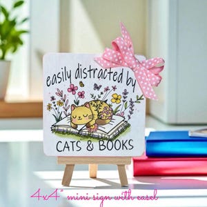 May include: A square white sign on a wooden easel featuring a cartoon cat sleeping on an open book surrounded by colorful flowers. The text reads "easily distracted by CATS & BOOKS". A pink and white polka dot ribbon is tied to the top corner.
