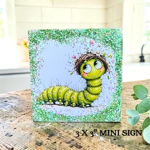 May include: A square 3 x 3 inch mini sign featuring a green caterpillar illustration with a small nest and flowers on its head. The artwork is framed by a border of green glitter and set against a light blue background with floral accents.