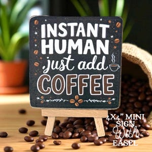 May include: A small, rectangular sign on a wooden easel. The sign is black with white and brown text that reads "INSTANT HUMAN just add COFFEE." Coffee beans and a coffee cup graphic are also on the sign. The sign is 4x4 inches.