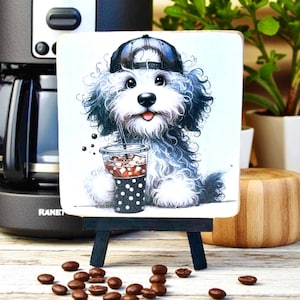 May include: A square wooden sign featuring a cartoon dog wearing a backwards baseball cap and drinking iced coffee. The dog is white and gray with a black cap and a black and white polka dot cup. Coffee beans are scattered in front of the sign.