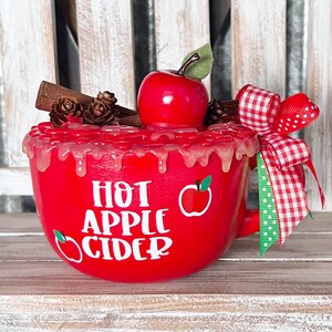 May include: A red mug featuring the white text HOT APPLE CIDER and two small apple graphics. The top is decorated with a faux red apple, cinnamon sticks, small pinecones, and a red and white checkered ribbon tied to the handle.