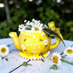 May include: A yellow teapot-shaped planter with white daisies and the text "Blooming Like A Daisy." The planter has a white scalloped trim and a yellow bow. Daisies are scattered around the planter on a white wooden surface.