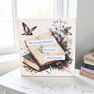 May include: A square wooden sign featuring an illustration of two stacked open books surrounded by red flowers and a bird. The text on the sign reads, My favorite character has ruined all real people. The design has a vintage, aged paper aesthetic.