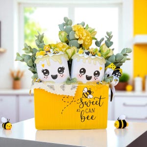 May include: A yellow container with the text "Sweet as can bee" holds two marshmallow-shaped figures with smiling faces, topped with yellow flowers and greenery. Decorative bees are placed around the container.