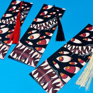 Può includere: Tre segnalibri neri con un motivo ripetuto di occhi bianchi con iridi rosse e una bocca con denti affilati. Ogni segnalibro ha un nappa rossa, nera o crema. Su sfondo blu.