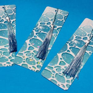 Puede incluir: Tres marcapáginas rectangulares con un diseño abstracto azul y blanco, cada uno adornado con una borla plateada. Los marcapáginas tienen un fondo azul y están decorados con un patrón de formas interconectadas y pequeñas chispas de colores.