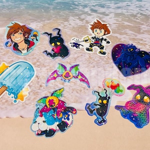 May include: A collection of colorful stickers featuring characters and designs from a video game. The stickers include a character with spiky hair, a popsicle, and various creatures. The stickers are on a sandy surface.