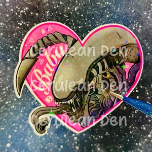 Op de afbeelding: Een hartvormige sticker met een beige xenomorph-wezen met een uitstekende binnenkaak en scherpe tanden. De achtergrond is felroze met kleine witte hart- en schedelpictogrammen. Het woord Baby is geschreven in wit schrift. De sticker heeft een holografische sterrenafwerking.