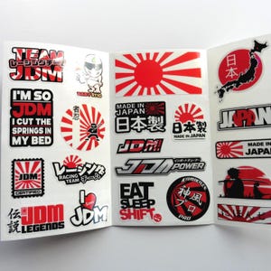 Op de afbeelding: Een verzameling JDM-stickers op een wit vel, met rode, zwarte en witte ontwerpen waaronder rijzende zon-motieven, Japanse tekst, autoslogans zoals 'Eat Sleep Shift' en diverse grafische illustraties van auto's en figuren.